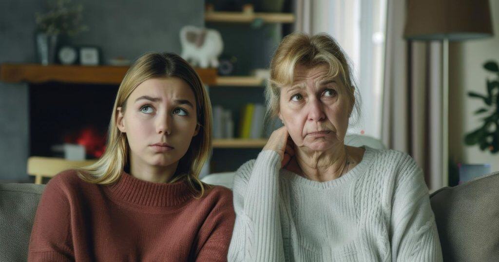 a mother and a daugher sitting next to each other on a couche in the living room looking awkward as they don't know what to say to each other after an argument.
