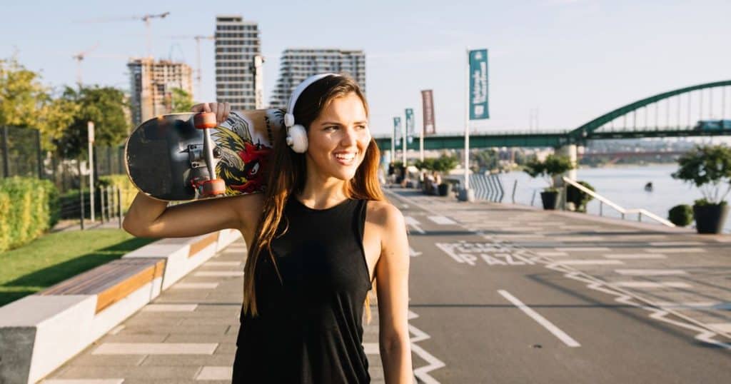 a young lady walking listening to music near a lakeside and carrying a skateboard on her shoulder feeling happy as her narcissistic parents praise her for being talented.