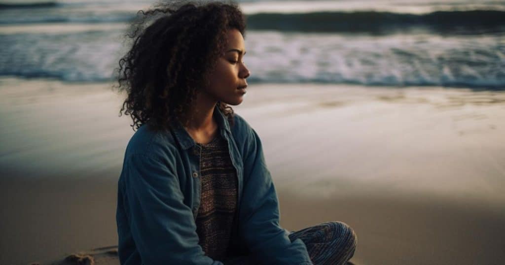 a woman wearing jean shirt sittong on the beach during sunset closing her eyes listening to soft wave breaks as she reflects on her experience with her narcissistic family.