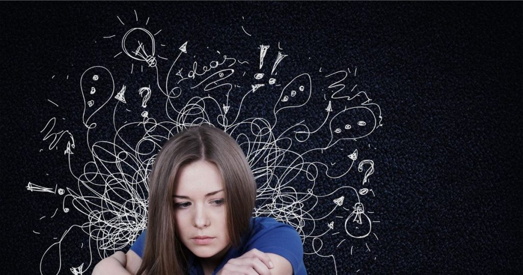 a woman with her long bronw hair wearing a blue t-shirt sitting alone against a black background with white random drawing looking sad.
