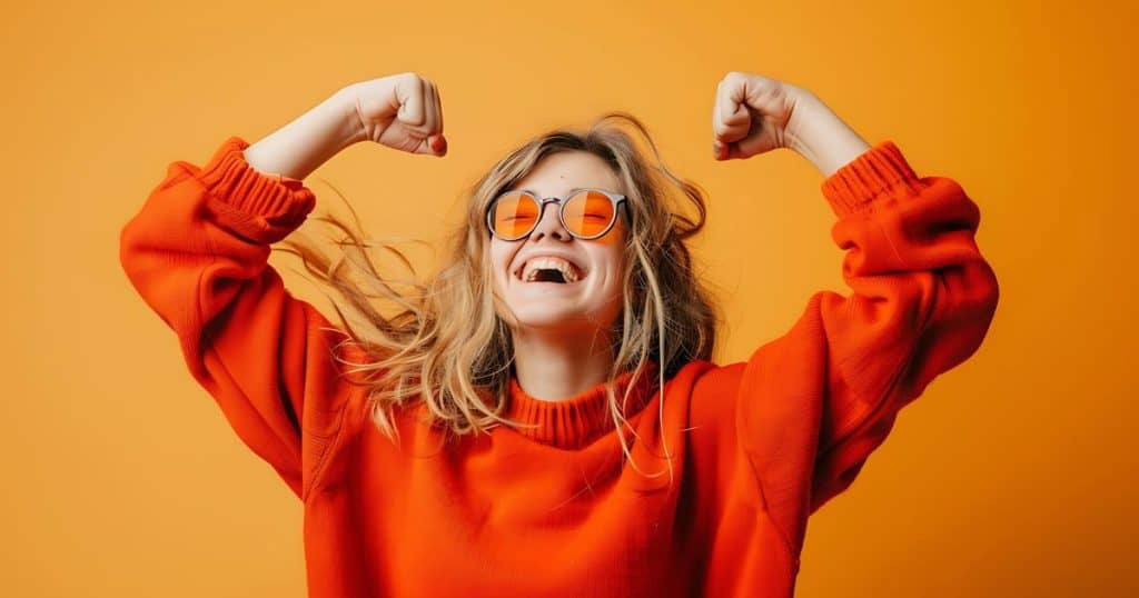 a woman in orange sweater, blonde hair and orange sunglasses with her arms flex smiling so big feeling victorious in a dark orange background.