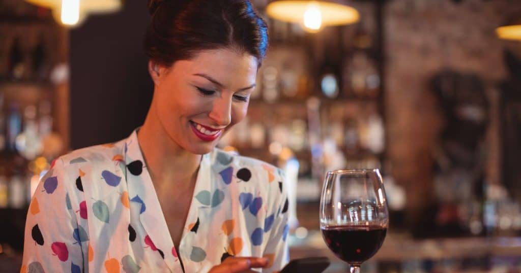 a beautiful woman with a clean updo and a white dress sitting in a bar with a glass of wine smiling while response to text messages from her toxic family's flying monkeys.
