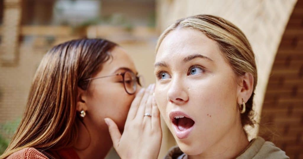 a close up shot of a woman with long brown hair wearing glasses is whispering into her girlfriend's ear a secret. Her girlfriend has her mouth wild open.