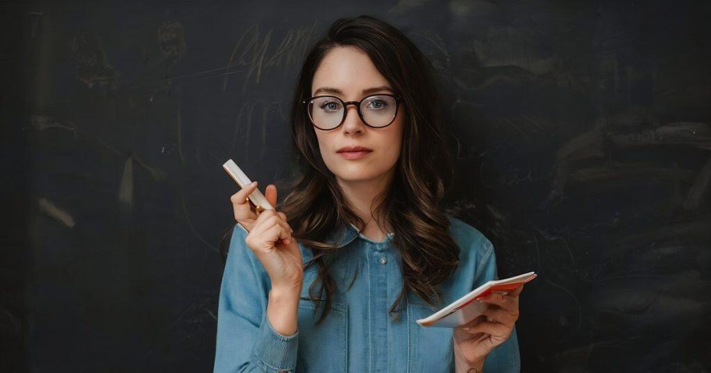a woman wearing glasses and jean shirt holding a chalk leaning against a dark board with a note in her hand about to teach on narcissistic family.