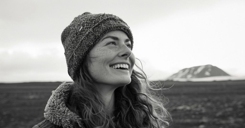 a black and white image a woman standing alone wearing a jacket and a hat during winter season smiling looking up in the sky.
