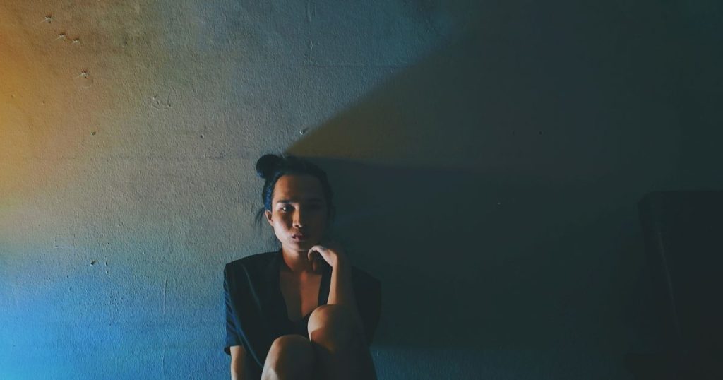 Portrait of young woman wearing an old navy dress with her hair tied up sitting against wall in darkroom alone looking confused and hopeless.