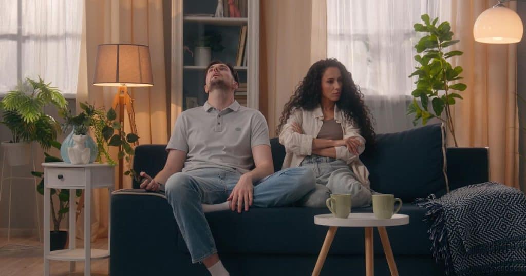 a woman and a man sitting next to each other on a blue couche in a living room well decorated. The woman is mad at her boyfriend as she has her arms crossed and her boyfriend has his head tilted back.