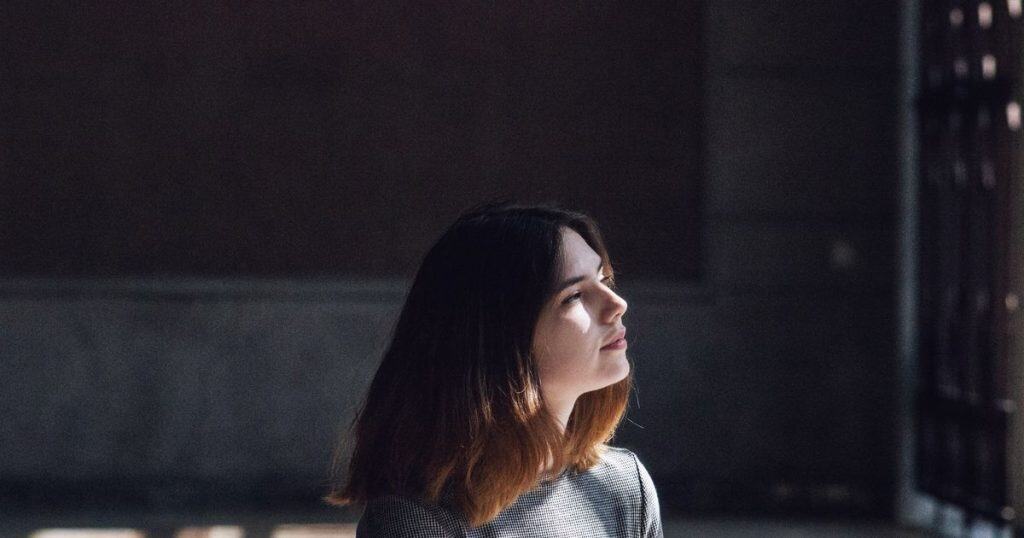 a woman with short black hair wearing grey t-shirt looking up at a sunny window in her living room looking peaceful.