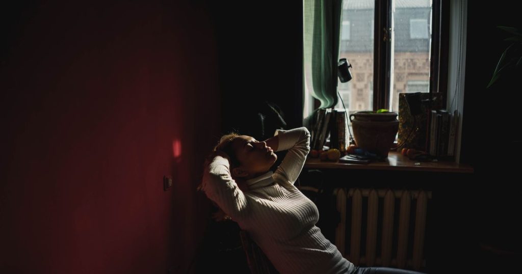 a woman wearing a white turtleneck sitting on a chair with her hands brushing her hair while closing her eyes in her red living room next to the window.
