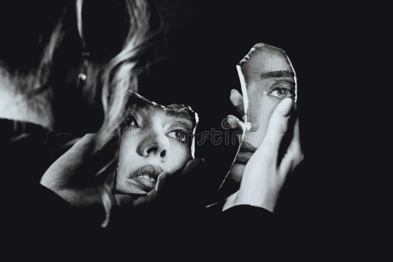 a black and white image of a woman holding a broken piece of mirror showing her reflection as she looks lost.