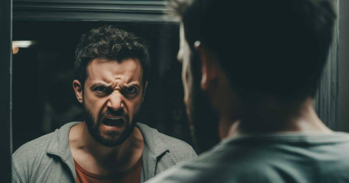 A distressed men with short dark brown hair is yelling into his bathroom mirror. His expression is angry and vulnerable, suggesting emotional tension.