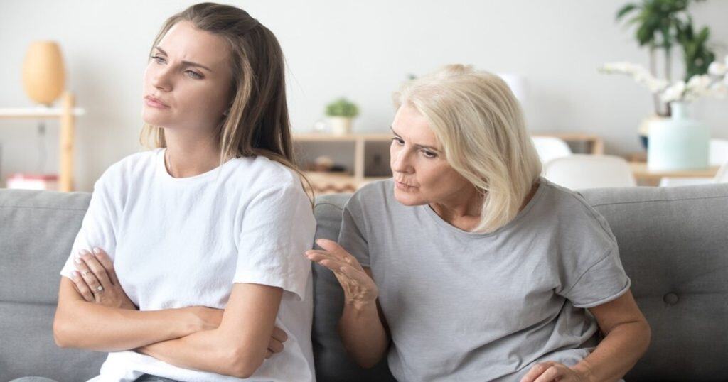 a mother and a daughter sitting on the couche in a white decor living room. The mother is trying to talk to her daughter who is upset with her.