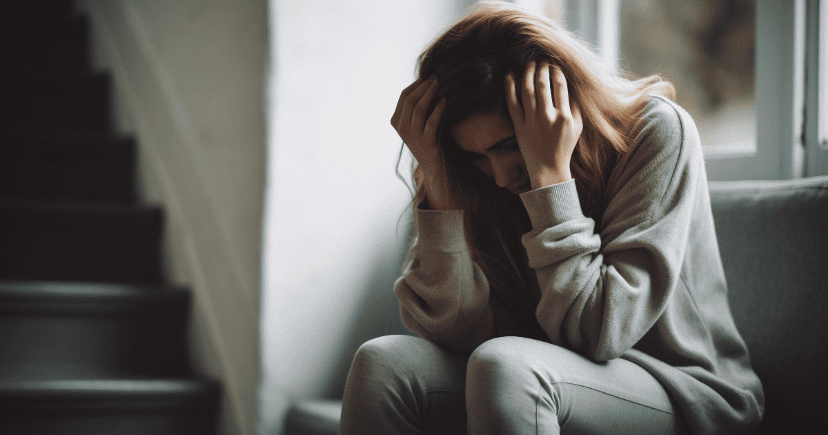 A woman sits on a couch near a window, hunched forward with her head in her hands, wearing a grey outfit, overwhelmed by emotion and tension in a dim, muted setting. It's as though manipulation wears the mask of love, guilt becomes the chain you mistake for your own choice.