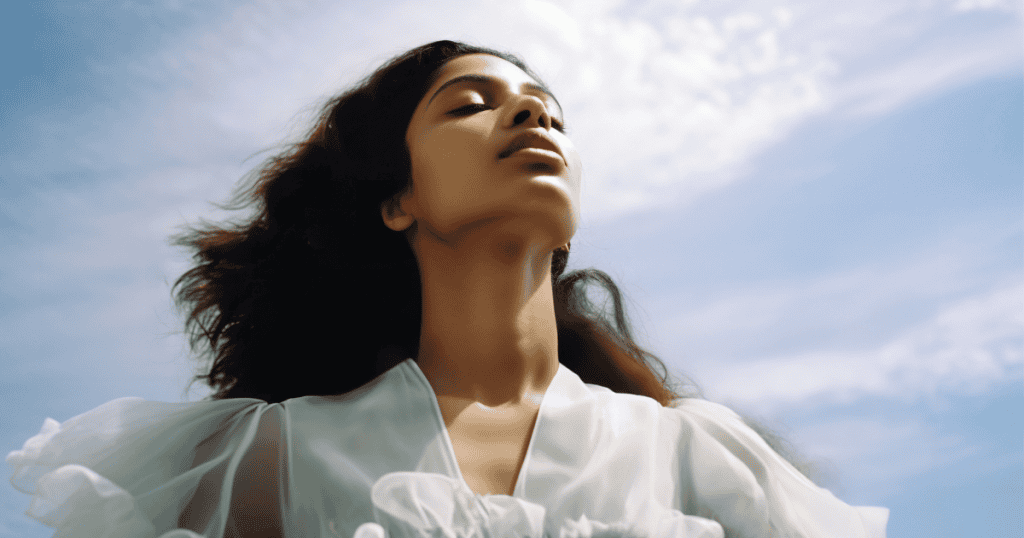 A serene young woman stands with her face lifted toward the sky, eyes closed in calm defiance, her white blouse flowing in the breeze under a clear sky. Her quiet strength is a reminder that no one gets to shrink your values and call it “being realistic.”