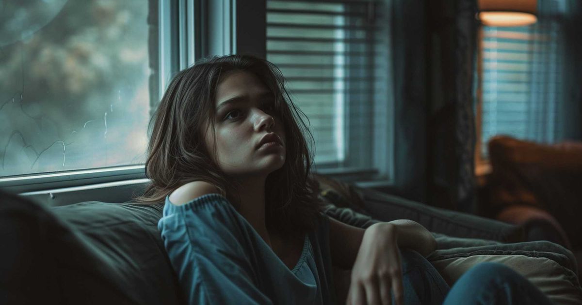 A young woman sits alone on a couch in a dimly lit room, staring pensively out the window. Her posture and expression convey emotional exhaustion and contemplation, reflecting the internal conflict of dealing with insincere apologies and unresolved tension in a toxic relationship.