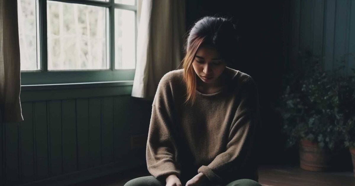 A young woman sits cross-legged on the floor in a dimly lit room, her head bowed and posture withdrawn, conveying a sense of loneliness and isolation as soft light streams in through a nearby window.