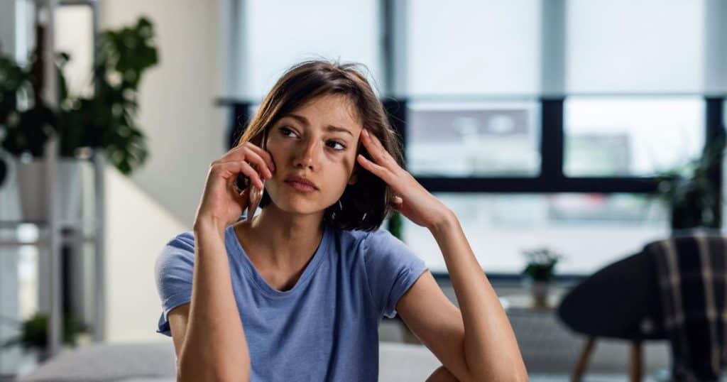 Young sad woman communicating over mobile phone and looking away while thinking of something sitting in her white decor lcondo living room.
