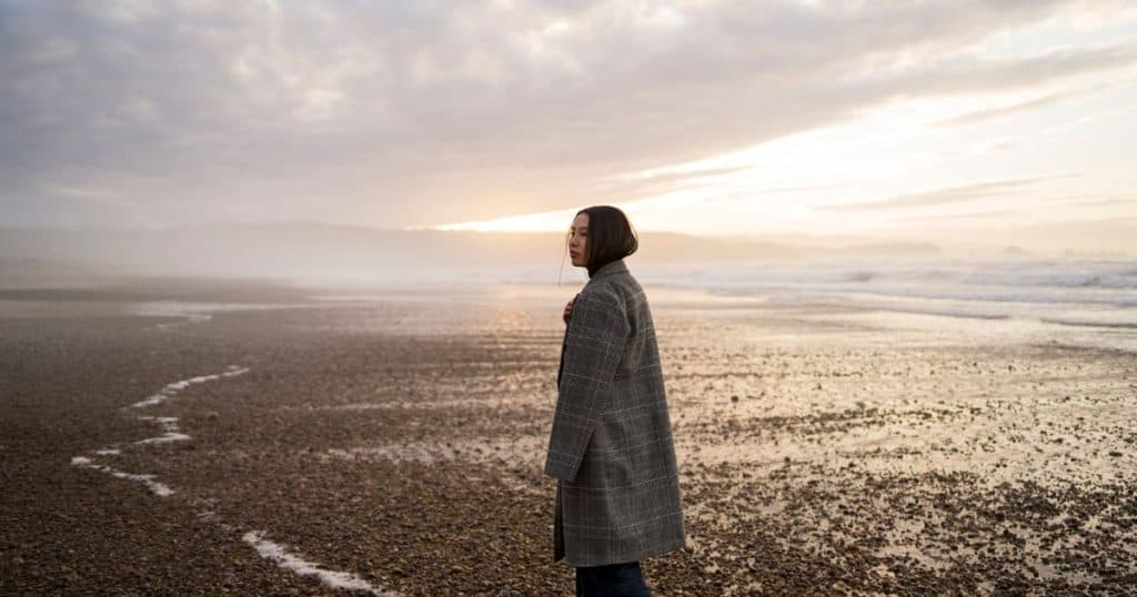 a woman in a trench grey coat is walking on the beach in the fall looking at the sunset feeling tense.