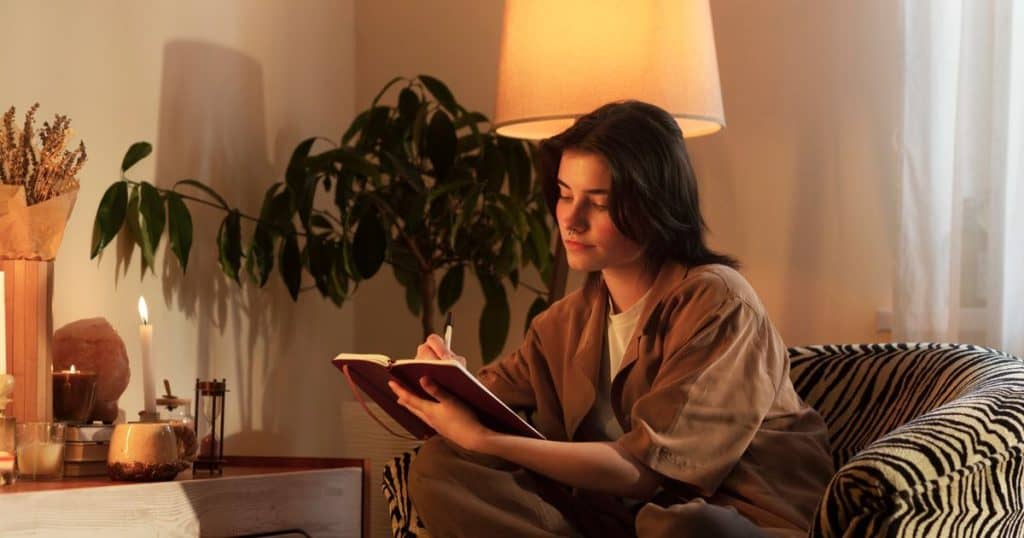 a woman with short black hair sitting in her cozy living room on a couche journaling away at night.