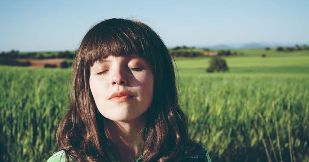 Woman with brown hair with her eyes closed standing on a green field during sunny day. Looking content and peaceful.