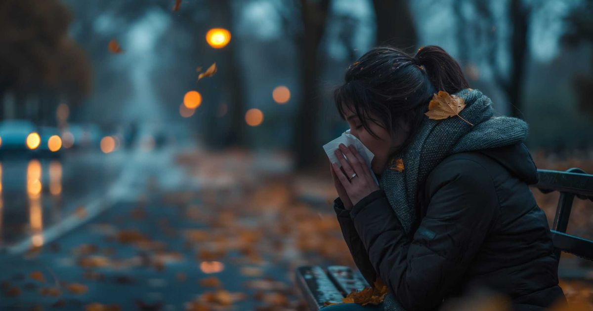 A woman sits alone on a park bench in the cold, autumn evening, bundled in a scarf and coat. She holds a tissue to her face, appearing tearful and emotionally overwhelmed. Fallen leaves surround her, and a few rest on her clothing, symbolizing the weight of emotional pain.