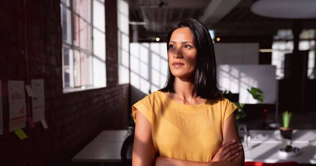 a dark hair woman wearing a yellow dress standing in her office smiling looking outside the window asa the sun shines on her face.