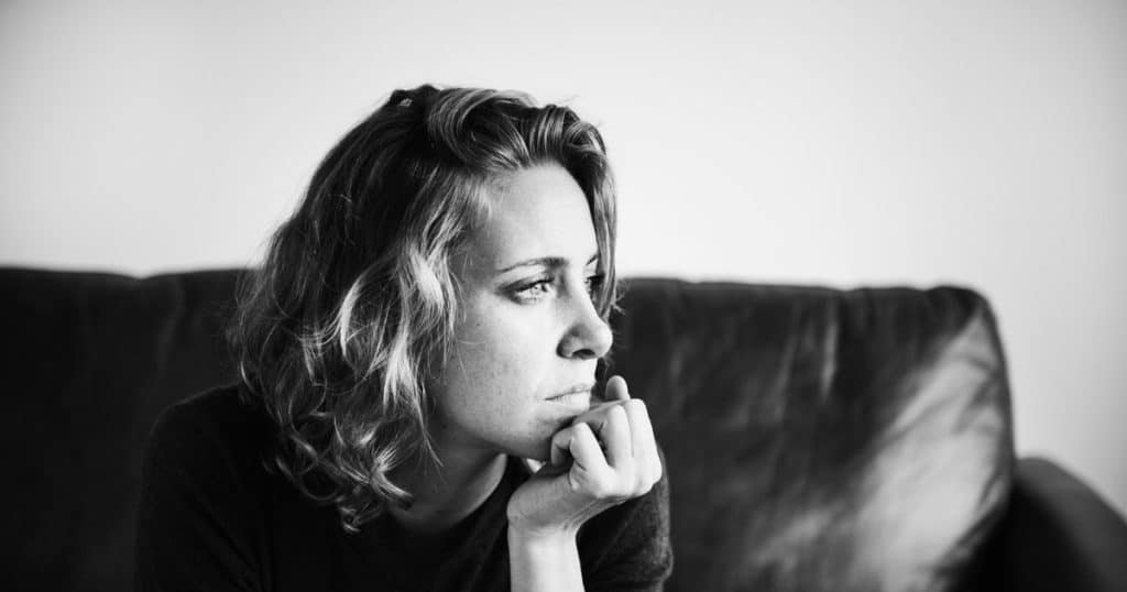 a black and white close up picture of a woman with short hair sitting in her black leather sofa looking outside her window feeling lost.