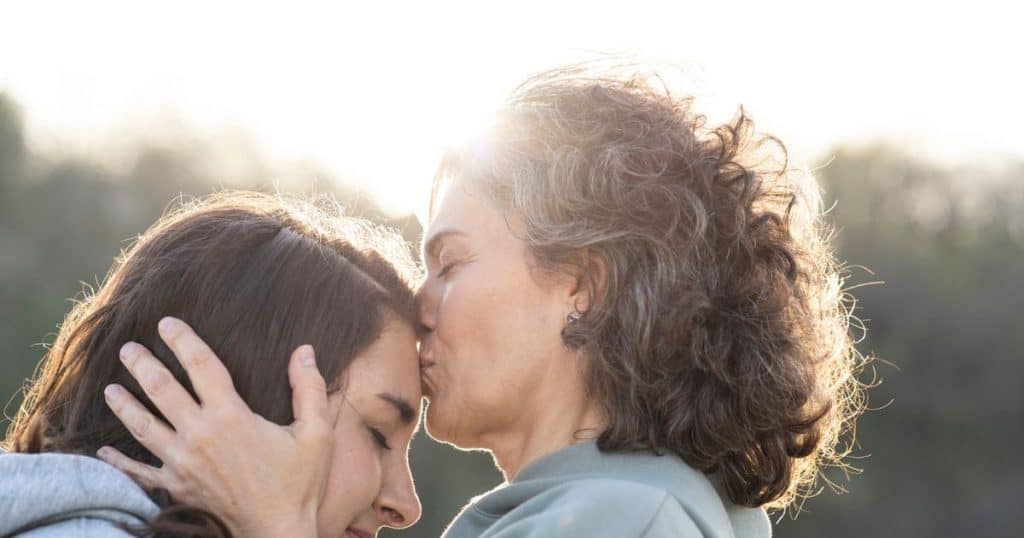 Mother kissing daughter's forehead both women standing out the park.
