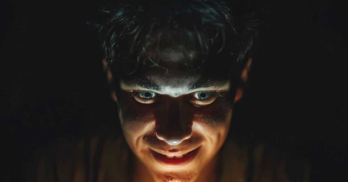 A dimly lit, intense close-up of a man with an eerie smile and glowing eyes, illuminated from below, creating a sinister and manipulative appearance.