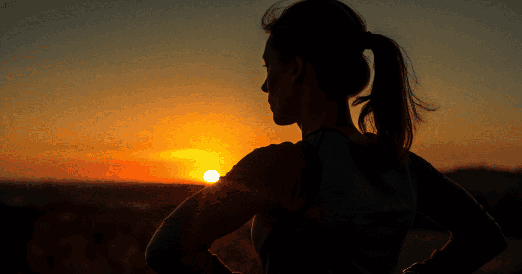 A woman stands in thoughtful silence with her hands on her hips, silhouetted against a vivid sunset, symbolizing reflection, realization, and inner strength. This explains that Sometimes, clarity only comes after darkness—when you finally name what broke you.