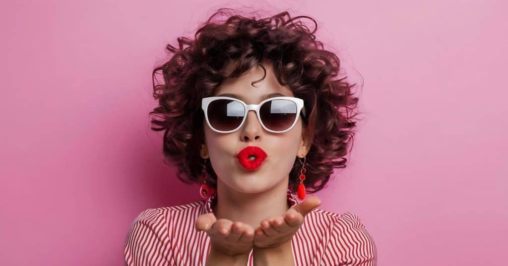 a curly hair woman with sunglasses and red lipstick blowing kisses standing against pink background looking fierce.
