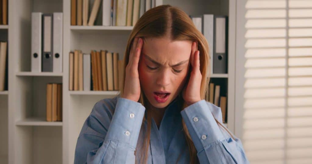 a woman wearing blue blouse standing in her office with her hands over her head as if she's having a headache looking angry.
