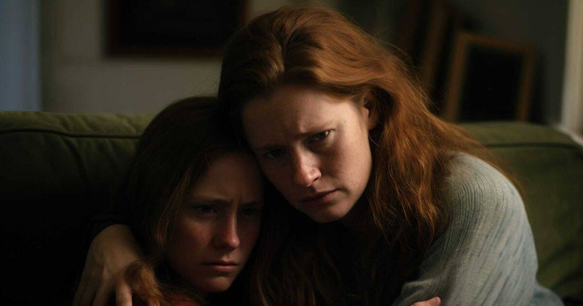 Two women with long red hair sit closely on a couch in a dimly lit room. One, appearing angry and protective, tightly embraces the other, who looks sad and vulnerable.