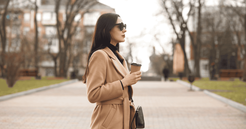 A woman in a camel coat and sunglasses holds a coffee cup while walking confidently, representing the strength of shutting down a narcissist without looking back.