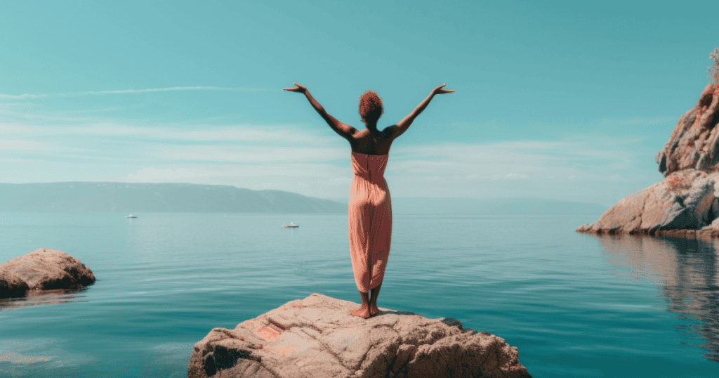 A woman in a pink dress stands on a rock with arms raised toward the ocean, symbolizing the freedom and strength that come from valuing self-worth over popularity.