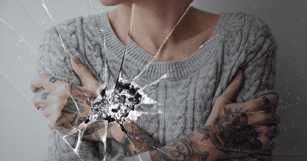 A person in a gray sweater with tattooed arms crossed over their chest is seen through cracked glass, symbolizing the restrictive and damaging rules narcissists impose.