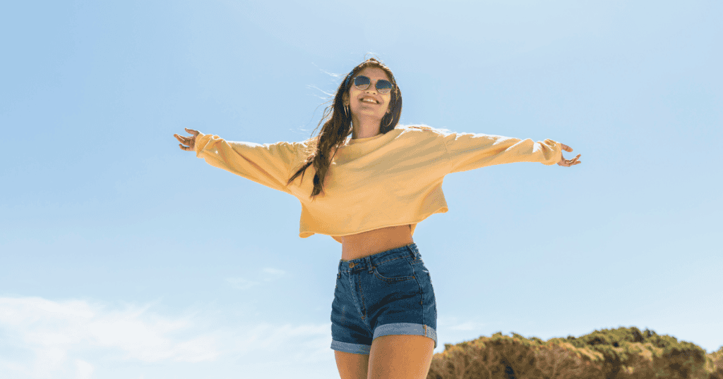 A smiling woman in sunglasses stretches her arms wide under a clear sky, symbolizing the freedom and empowerment of no longer being controlled by a narcissist.