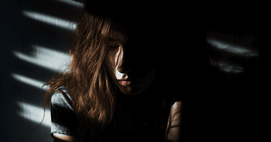 A young woman sits in partial darkness with shadows across her face, reflecting how narcissists teach you to hide your needs.
