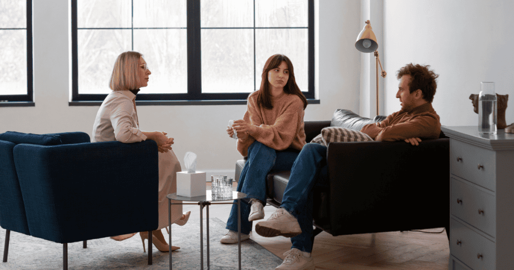 A woman sits between a man and another woman in a therapy session, symbolizing the process of examining family opinions and expectations to see if they align with her true self.