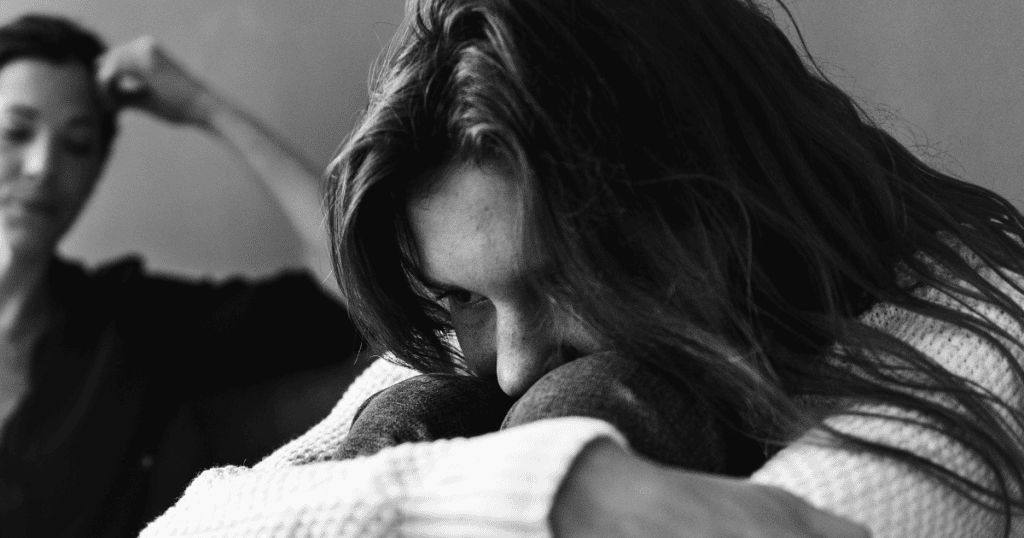 Black-and-white image of a distressed young woman sitting with her head on her knees while another person looms in the background, symbolizing the emotional burden placed on the scapegoat.