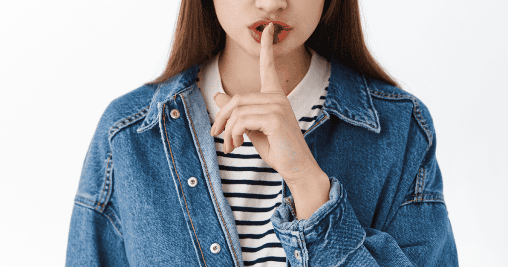 A woman in a denim jacket holds a finger to her lips in a shushing gesture, symbolizing the truths narcissists will never openly admit.