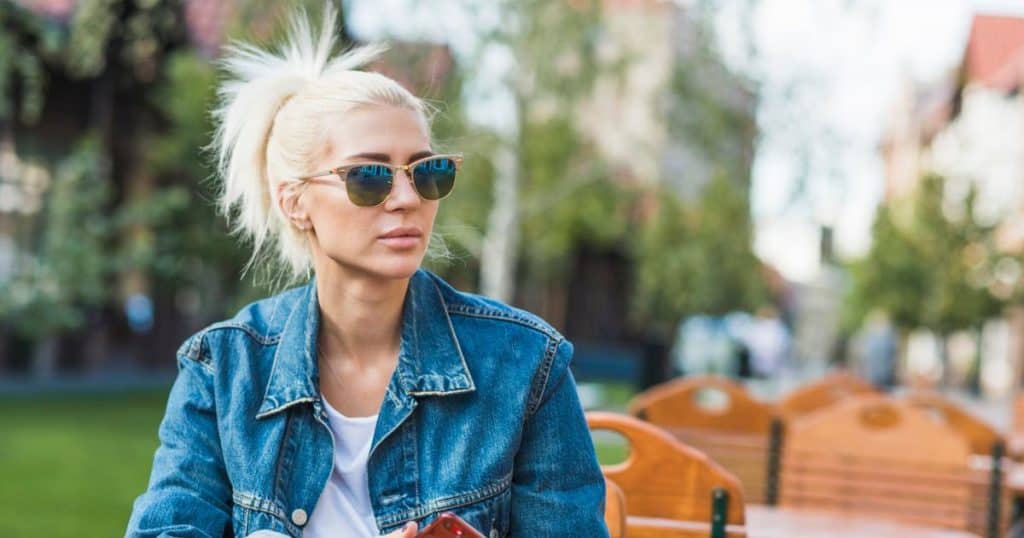 a cool blond woman with sunglasses on holding her phone looking firm and confident sitting outside the restaurant's patio.
