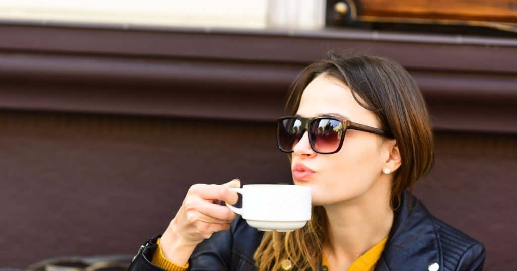Girl in sunglasses drinks tasty coffee on brown terrace background, defocused. Woman with busy face spends time in cafe. Hot beverage and break concept. Lady has cup of espresso during coffee break