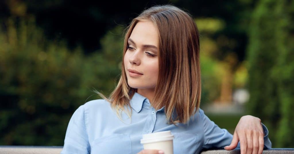 a woman with short brown hair sitting outside wearing blue blouse with smirk on her facing holding a cup of coffee.