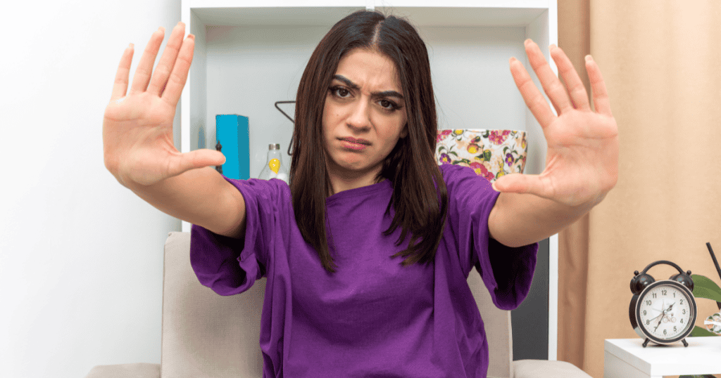 A woman sitting indoors raises both hands in a firm stop gesture, showing how to set boundaries with a narcissist who targets you.