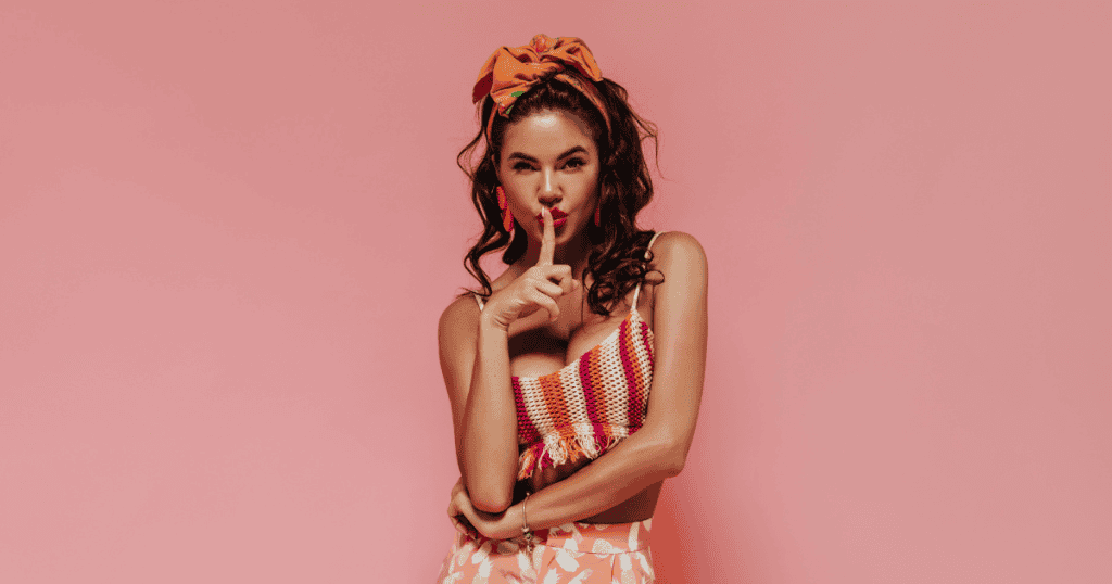 A confident woman stands against a pink background with her finger held to her lips in a hush gesture, symbolizing the firm rules she follows every time a narcissist crosses her mind.