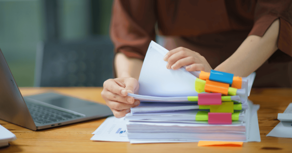 A person organizes a stack of papers with colorful tabs beside a laptop, symbolizing the importance of documenting everything to outsmart a narcissistic boss.