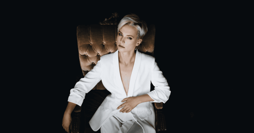 A woman in a white suit sits confidently in a chair against a dark background, symbolizing entitlement as the driving thread behind narcissism.