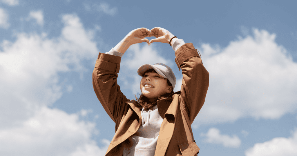 A woman smiles and makes a heart shape with her hands under the sky, showing that healing after narcissistic grief can still hold space for love.