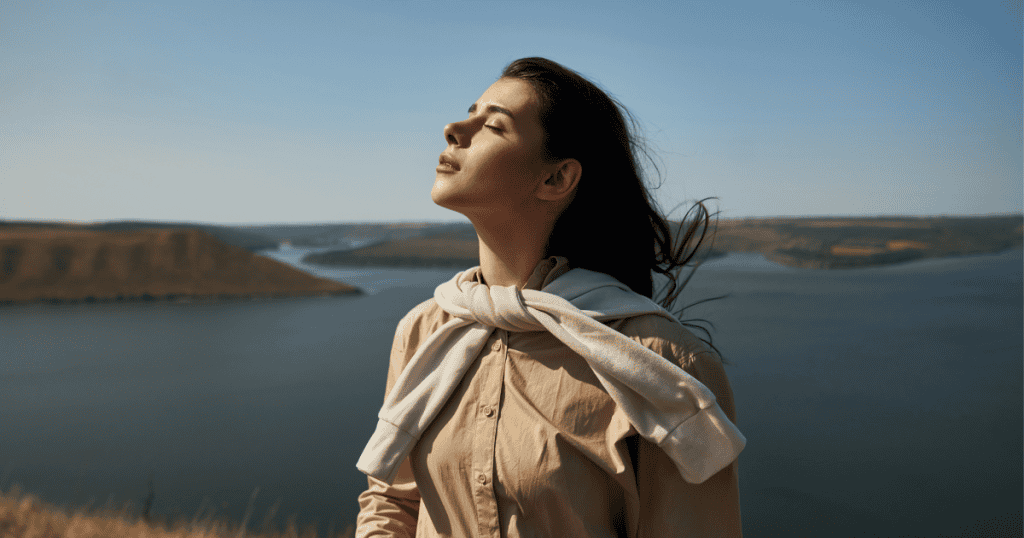 A woman stands peacefully by the water with her eyes closed and face lifted to the sun, symbolizing how knowing the faces of narcissism shifts your power toward freedom and clarity.